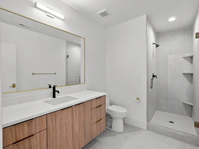 Wood-finish vanity with a white countertop and undermount sink, large mirror with a brass-finish frame, and walk-in shower with tiled walls and glass enclosure