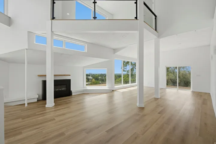 Open-concept living space featuring high ceilings, wood-finish flooring, a contemporary fireplace with a wood mantle, an upper-level balcony with a glass railing, and multiple large windows