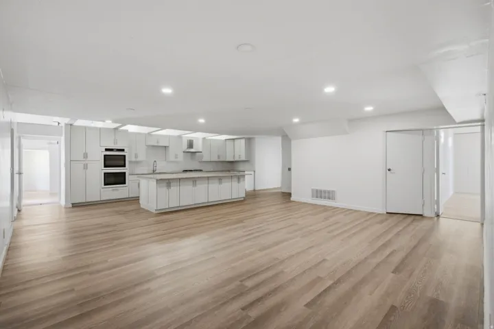 Open-concept living area with wood-finish flooring, recessed lighting, and a kitchen featuring white cabinetry, an island, and built-in double ovens
