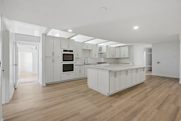 Kitchen featuring white shaker cabinetry, double wall ovens, a large island with a light-toned countertop, a subway tile backsplash, and wood-finish flooring