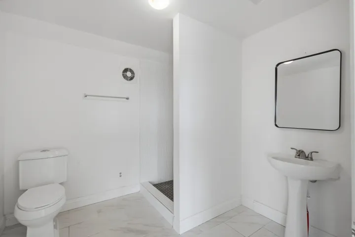 Pedestal sink with a framed mirror, white toilet, and a shower area with a ventilation fan