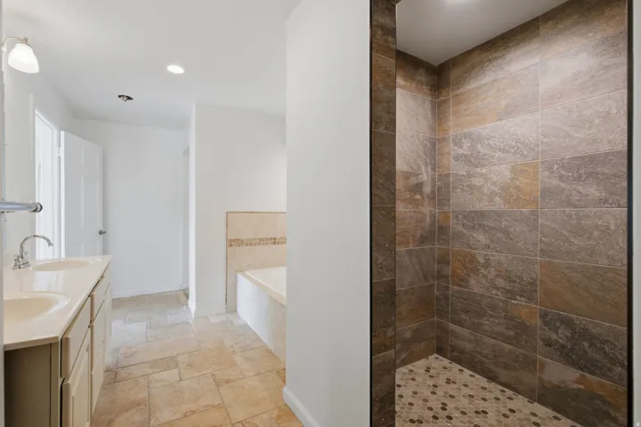 Spacious bathroom featuring a dual vanity, built-in bathtub with tiled surround, and a walk-in shower with stone-look tile walls and a mosaic tile floor