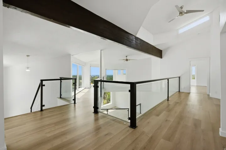 Open-concept interior featuring high vaulted ceilings, light wood-finish flooring, glass and black metal railing, and a prominent exposed beam