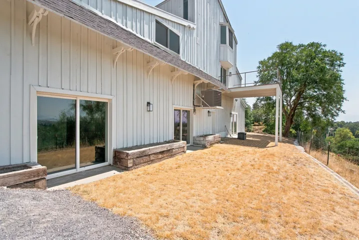 Board and batten siding, multiple sliding glass doors, upper-level balcony, covered patio area, and exterior wall sconces