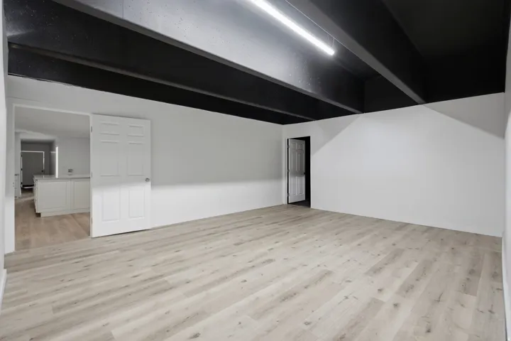 Spacious room featuring light wood-finish flooring, white walls, and a black exposed beam ceiling with track lighting