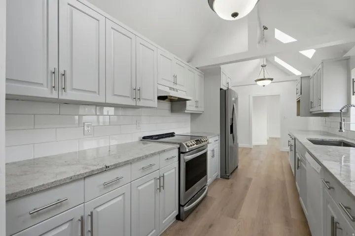 Vaulted ceiling with skylights, light wood-finish flooring, white shaker-style cabinetry, stainless steel appliances, and subway tile backsplash