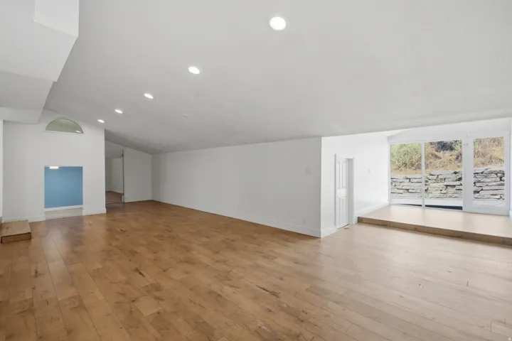 Expansive space with wood-finish flooring, recessed lighting, white walls, a stepped area with glass doors, and a decorative arched window