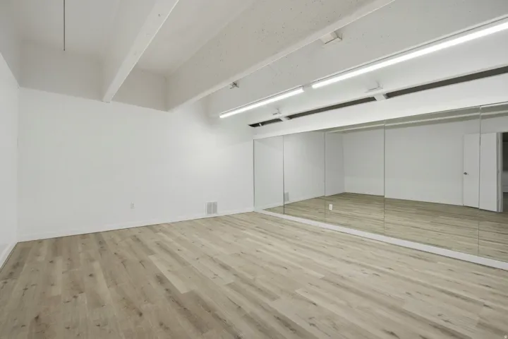 Expansive room with wood-finish flooring, white walls, exposed ceiling beams, fluorescent lighting, and a mirrored wall