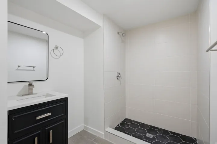 Bathroom featuring a dark wood vanity with a white countertop, a rectangular mirror, a shower with white tile walls, and a black hexagonal tile shower floor