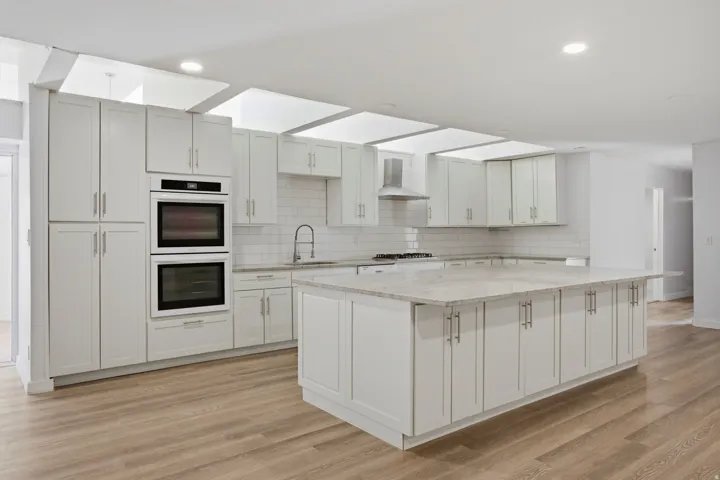 Kitchen featuring light wood-finish flooring, white cabinetry, a large kitchen island with light-toned countertops, and built-in double ovens