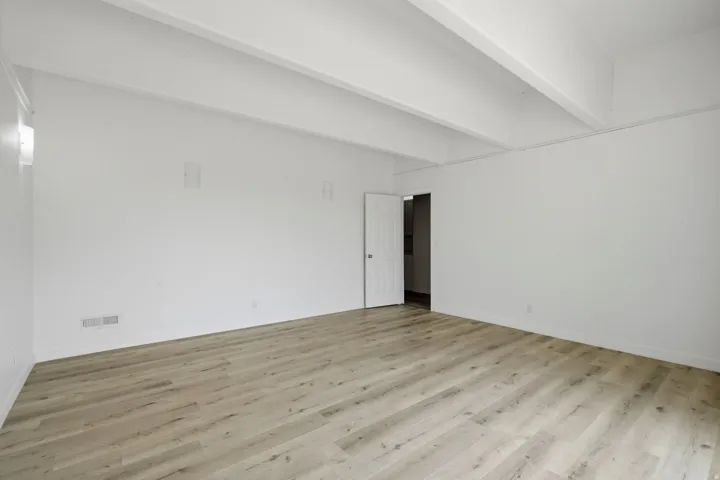 Spacious room featuring wood-finish flooring, white walls, and exposed ceiling beams