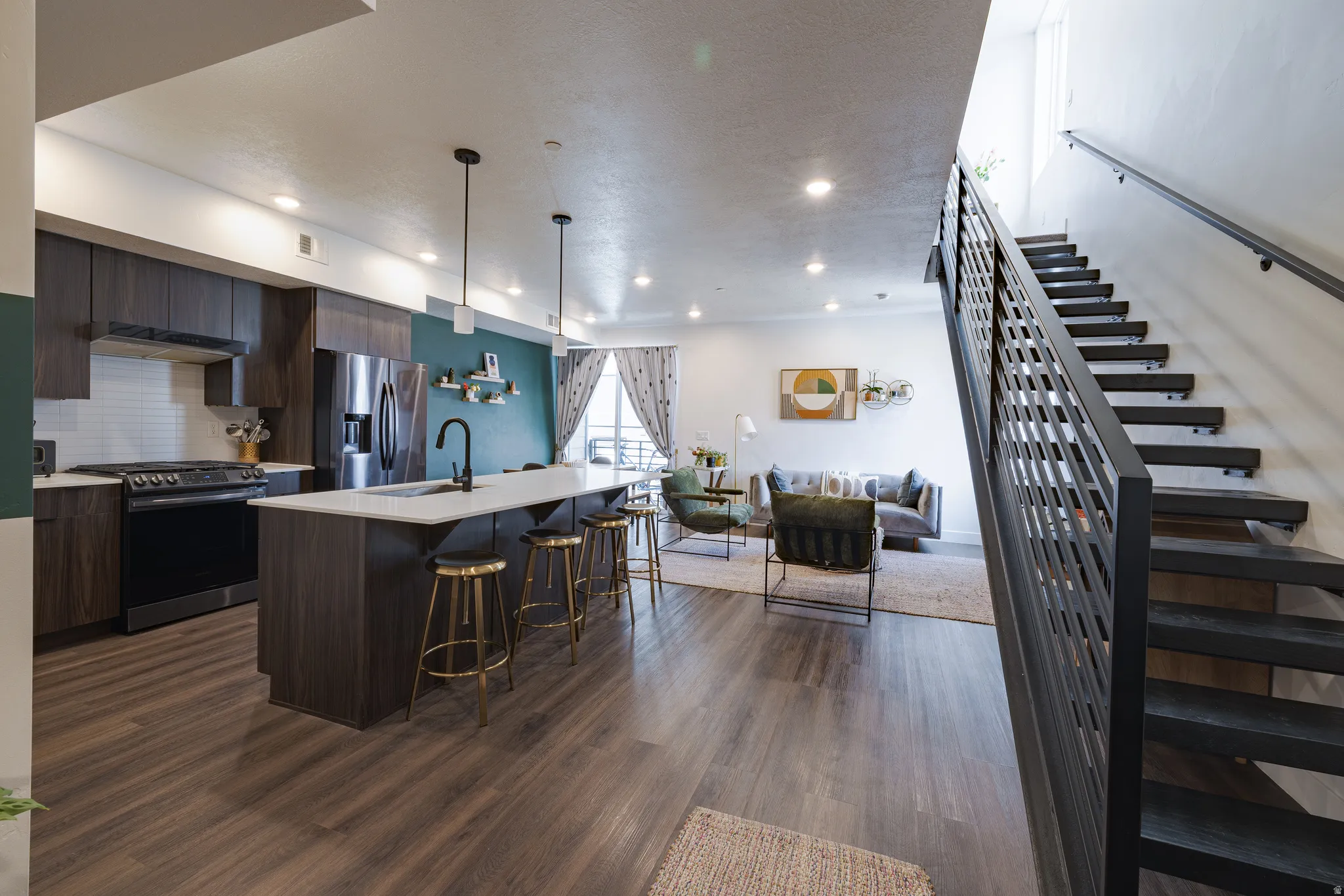 Open-concept living area with wood-finish flooring, a contemporary kitchen, a kitchen island with seating, a modern staircase with a metal railing, and recessed lighting
