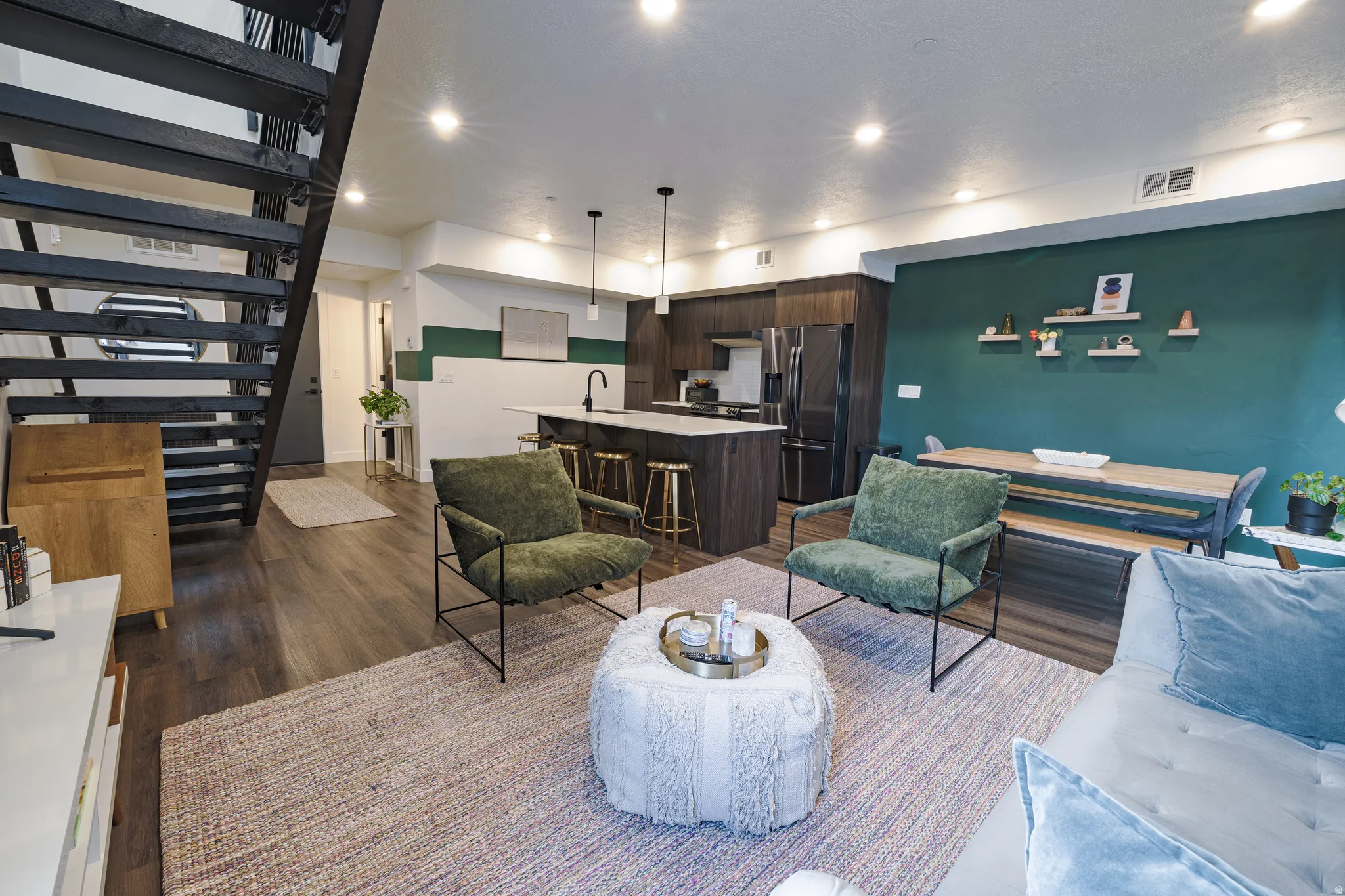 Open-concept living space featuring wood-finish flooring, a modern kitchen with dark cabinetry and a center island, recessed ceiling lighting, and an open-tread staircase with black railing