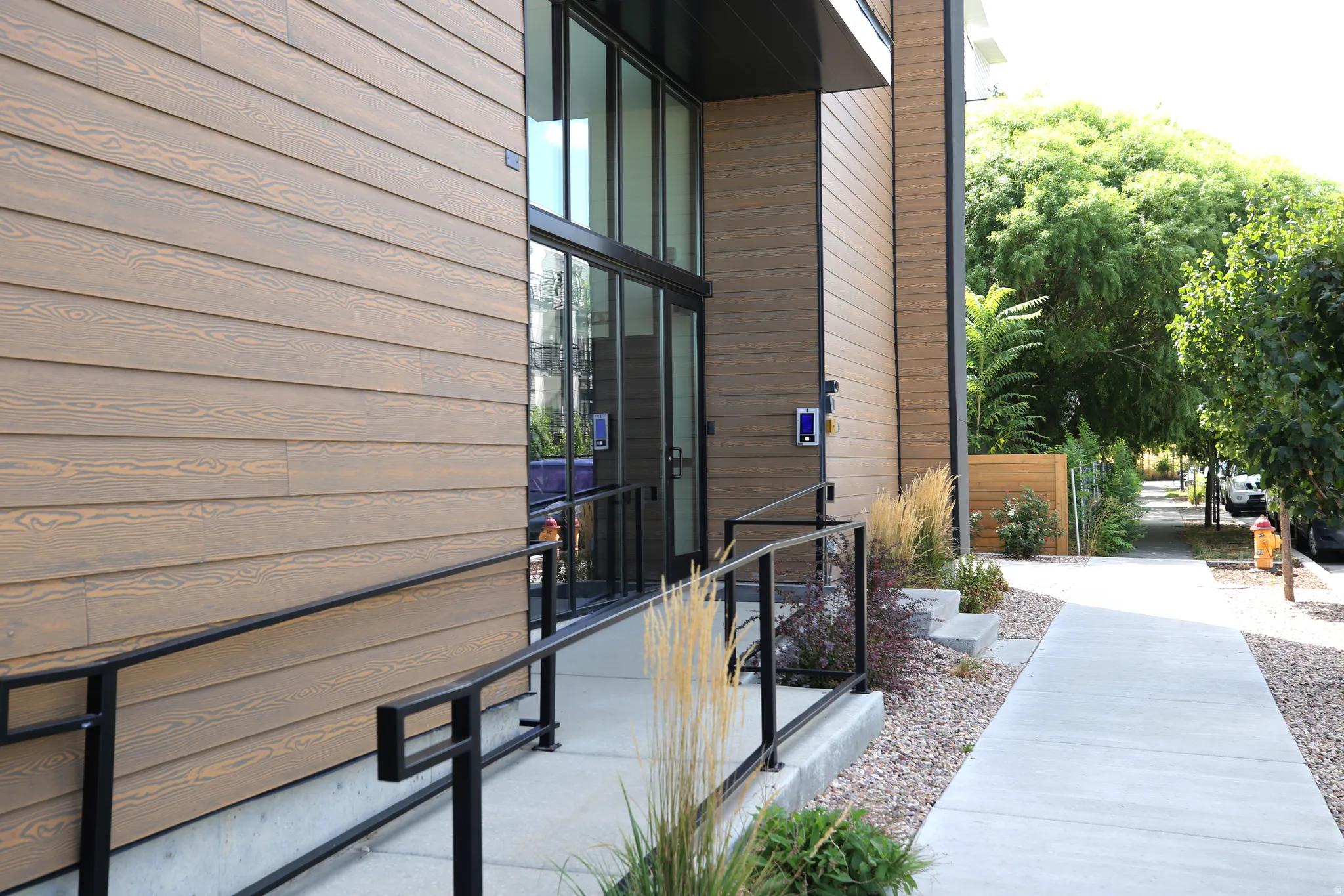 Modern exterior featuring wood-finish siding, full-light entry doors, and a concrete ramp with black metal railings; secure entry requires a digital key.