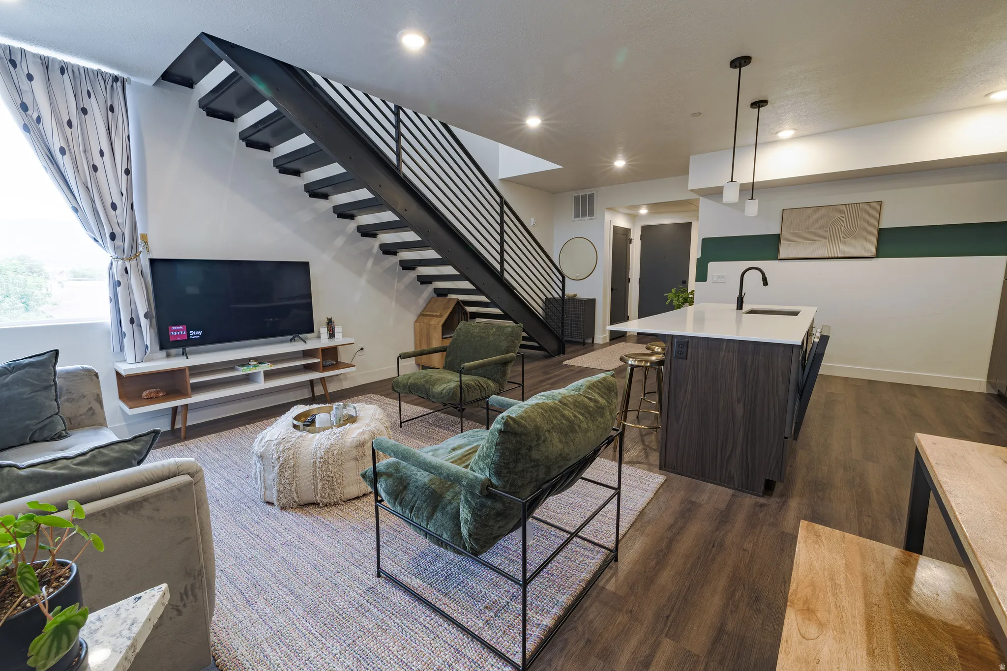 Open-concept living space with dark wood-finish flooring, a contemporary floating staircase with metal railings, a kitchen island with a light-colored countertop and integrated sink, and recessed lighting