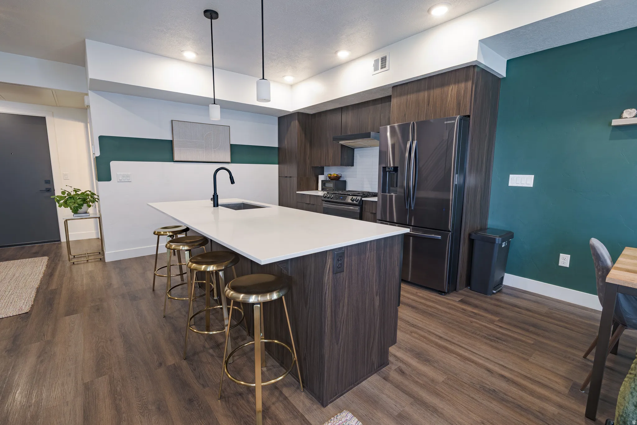 Modern kitchen featuring a large waterfall-edge island with white quartz countertop, dark wood cabinetry, stainless steel appliances including a French-door refrigerator, matte black faucet with pull-down sprayer, pendant lighting, and wood-finish floorin