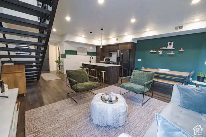 Open-concept living space featuring wood-finish flooring, a modern kitchen with dark cabinetry and a center island, recessed ceiling lighting, and an open-tread staircase with black railing