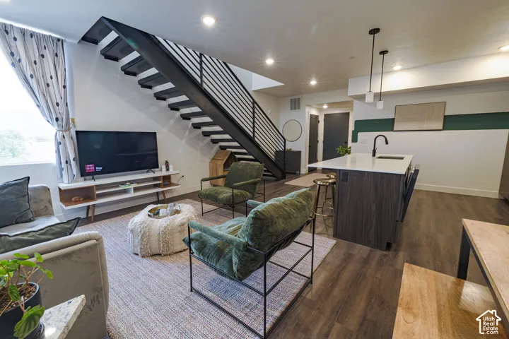 Open-concept living space with dark wood-finish flooring, a contemporary floating staircase with metal railings, a kitchen island with a light-colored countertop and integrated sink, and recessed lighting