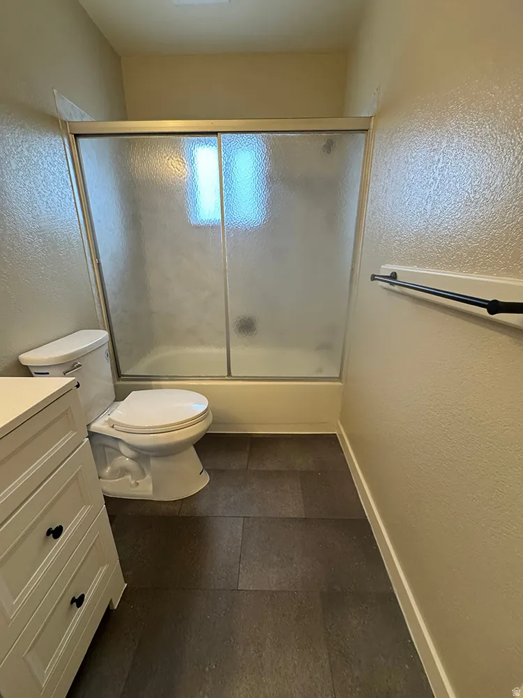 Bathtub with sliding frosted glass enclosure, white vanity with black hardware, and dark tile flooring