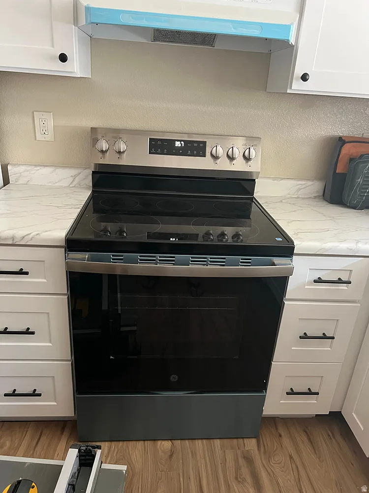 Stainless steel range with a glass cooktop, white shaker-style cabinetry, matte black hardware, white countertops with veining, and wood-finish flooring