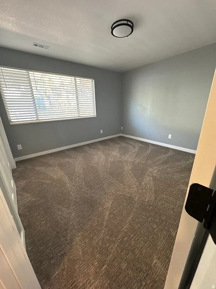 Neutral gray wall paint, brown patterned carpet, window with horizontal blinds, and a flush mount ceiling light