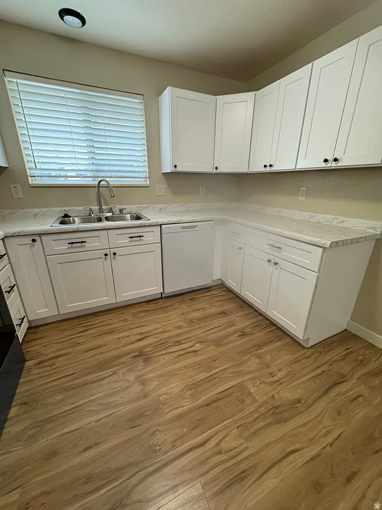 White shaker-style cabinetry, light-toned countertops, stainless steel sink, wood-finish flooring, and a built-in dishwasher