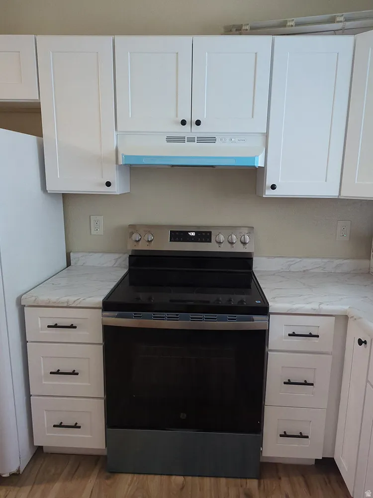 White shaker-style cabinetry with black hardware, stainless steel range, white range hood, light-toned countertops, and wood-finish flooring