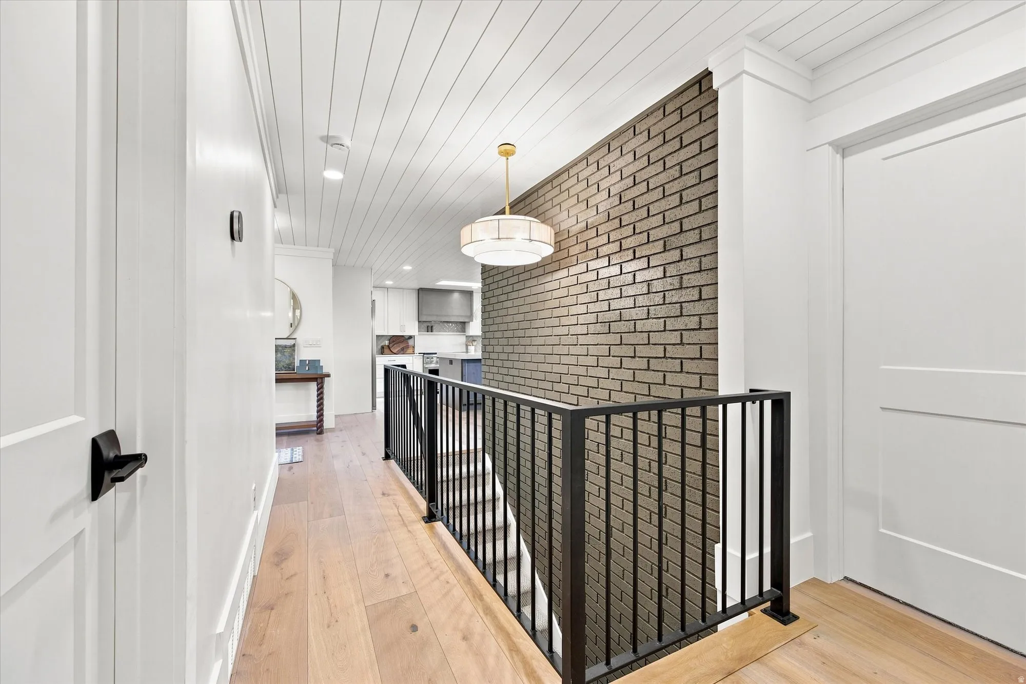 Wide plank wood-finish flooring, white shiplap ceiling, contemporary chandelier, exposed brick accent wall, and a black metal staircase railing