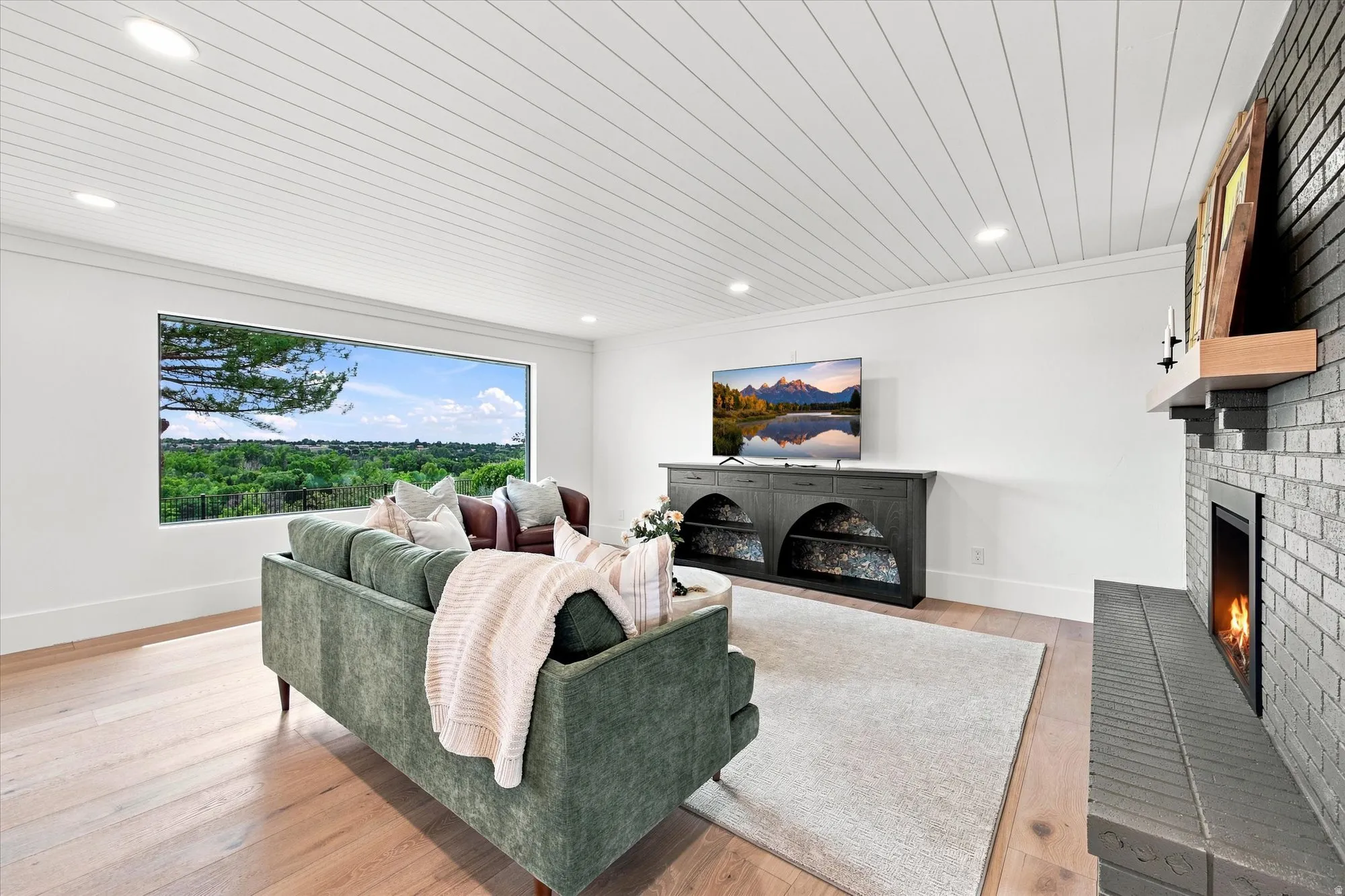 Wide-plank wood-finish flooring, white shiplap ceiling with recessed lighting, whitewashed brick fireplace with wood mantel, and a built-in media console