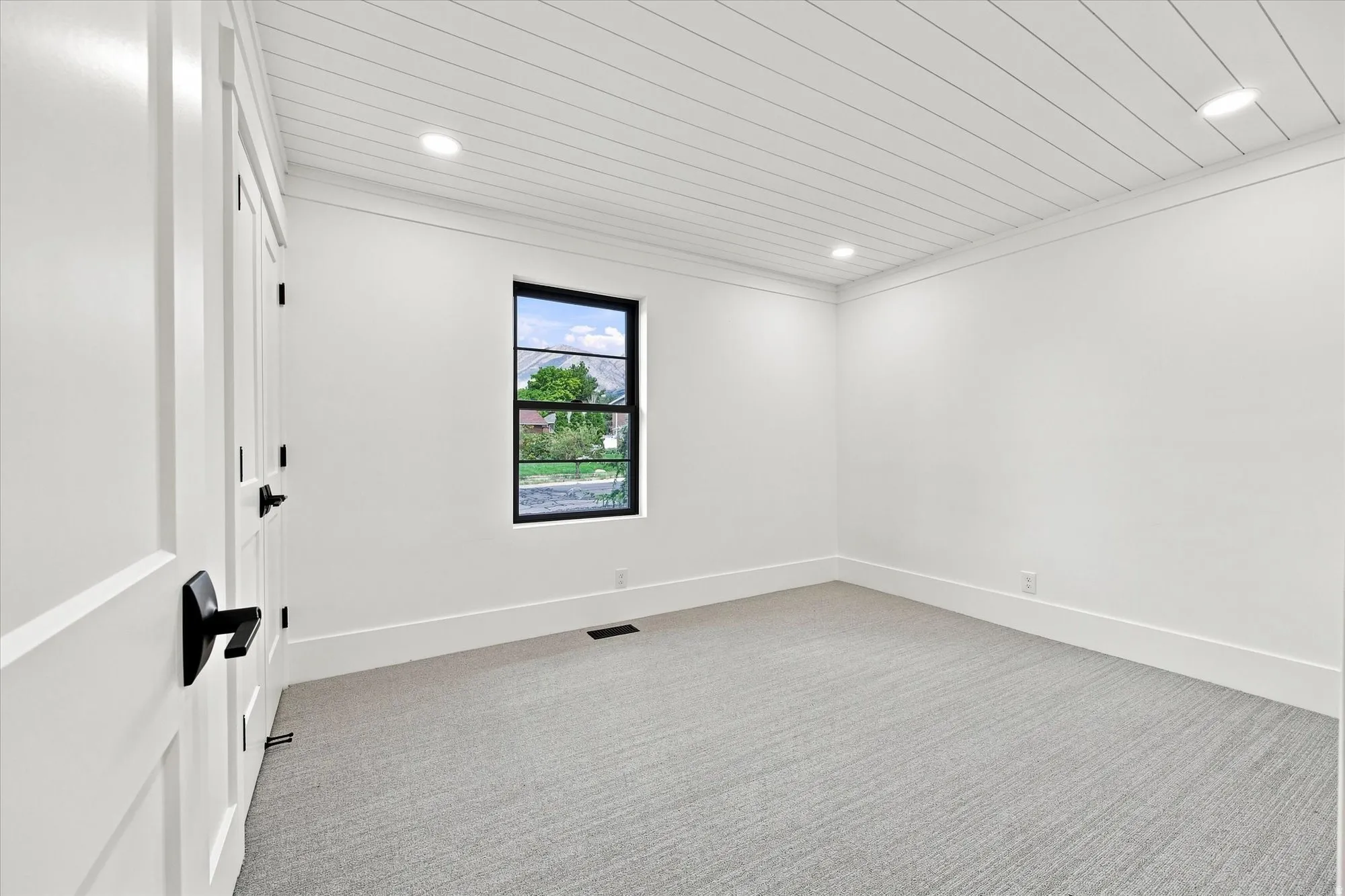 White paneled ceiling with recessed lighting, single window with black trim, gray patterned carpet, and white paneled doors with matte black hardware