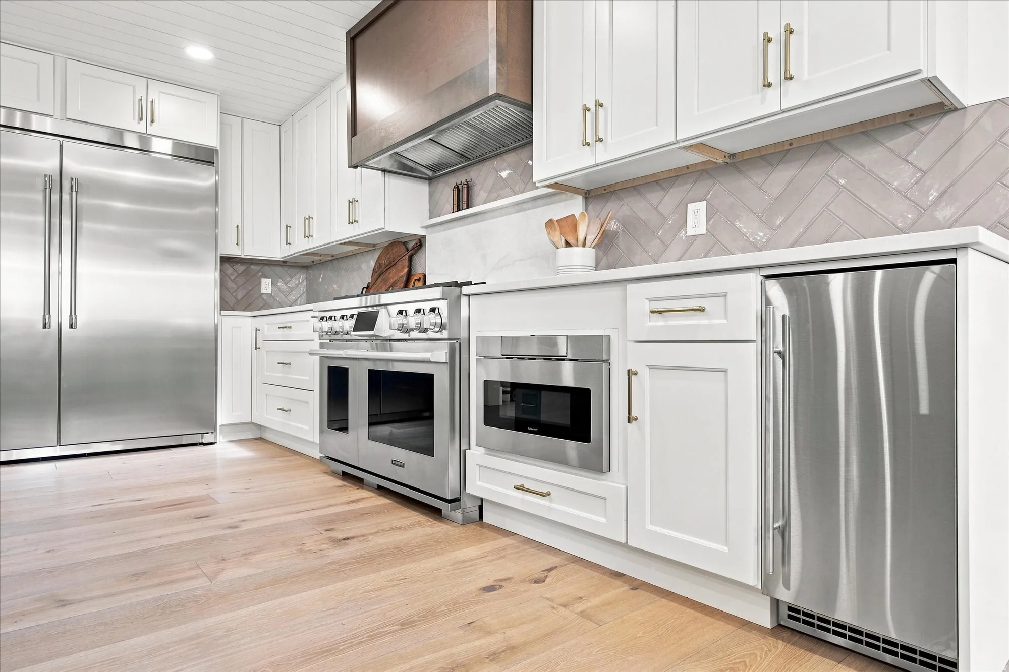 Stainless steel appliances, white cabinetry with gold-finish hardware, herringbone tile backsplash, white countertops, and wood-finish flooring