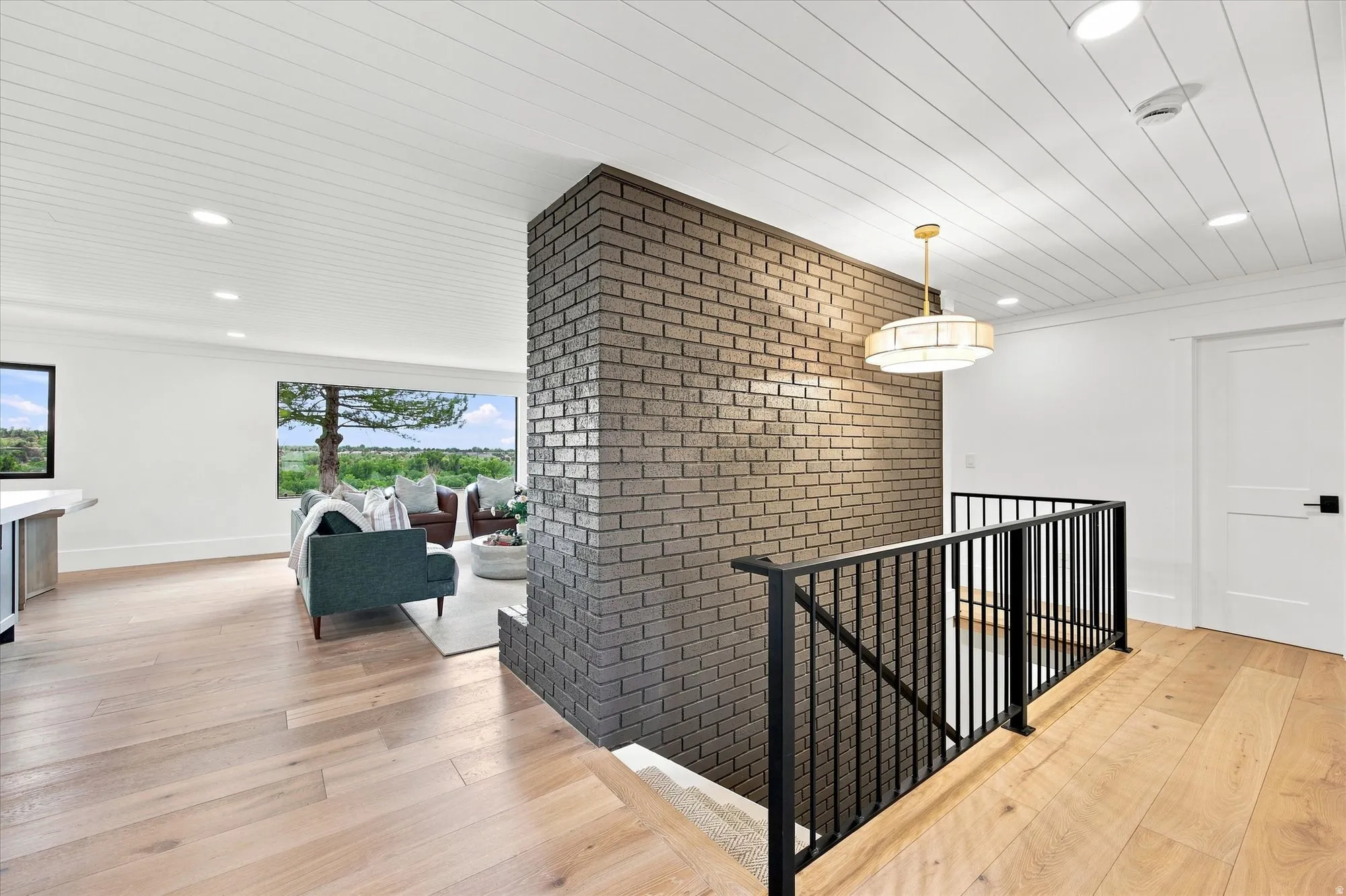 Wide plank wood-finish flooring, white shiplap ceiling, recessed lighting, exposed brick accent wall, and matte black stair railing