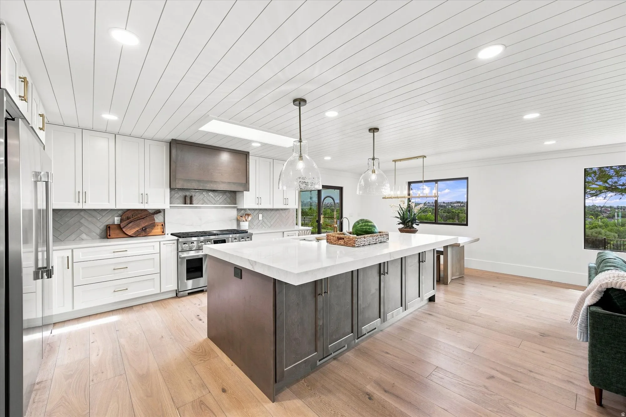 Gourmet kitchen with a large central island, white shaker cabinetry, stainless steel appliances, herringbone tile backsplash, and light wood-finish flooring