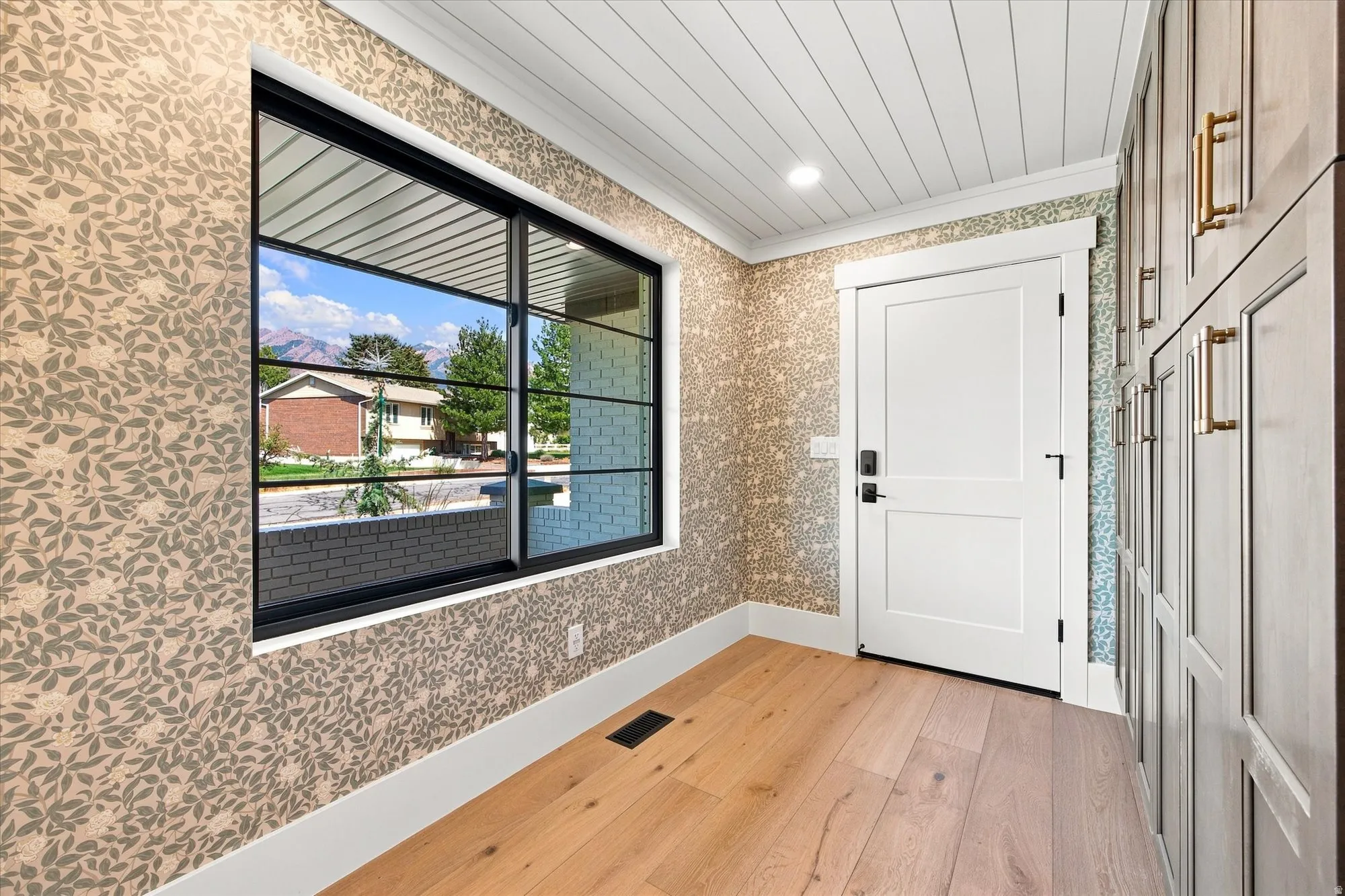 Welcoming entryway featuring warm wood-finish flooring, textured wallcovering, white shiplap ceiling, and a large black-framed window with natural light