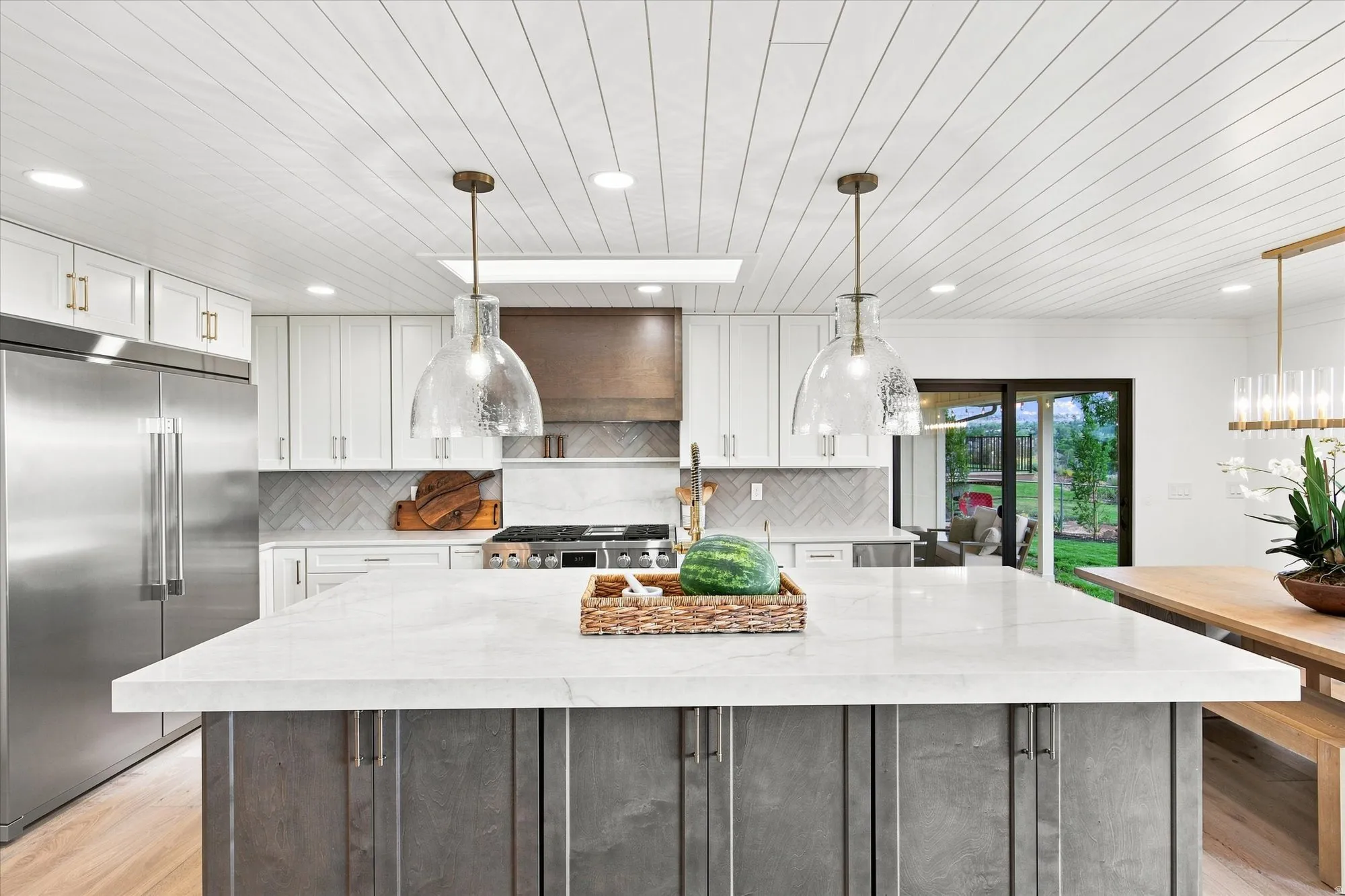Center island with a light-toned countertop, pendant lighting, stainless steel refrigerator, white cabinetry with gold-toned hardware, and a wood-finish ceiling