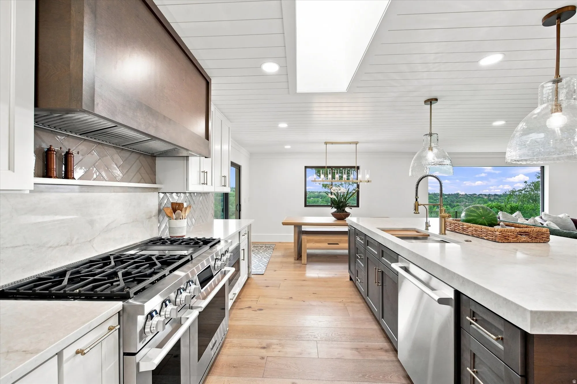 Gourmet kitchen featuring a stainless steel range, white shaker cabinetry, an island with an integrated sink, light wood-finish flooring, and a rectangular skylight