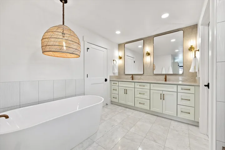 Freestanding soaking tub with a bronze-finish faucet, dual vanity with light-colored cabinetry, stone-finish countertop, and two rectangular wall-mounted mirrors