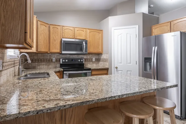 Kitchen featuring L-shaped granite countertops, wood-finish cabinetry, stainless steel appliances, a tile backsplash, and a stainless steel sink with a gooseneck faucet