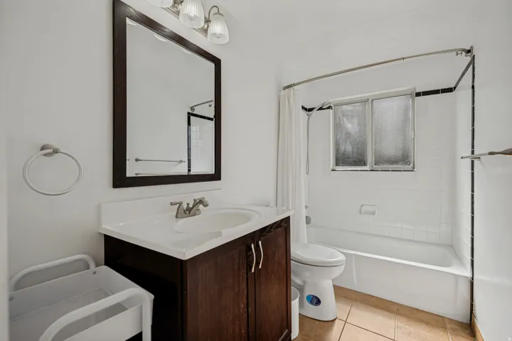 Vanity with a white countertop and integrated sink, framed mirror, wall-mounted light fixture, and a tiled shower-tub combination with a window