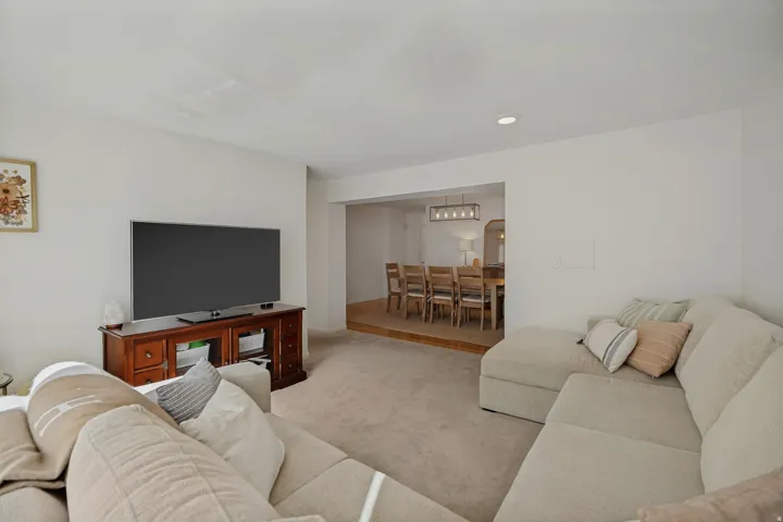 Carpeted living area with recessed lighting, offering access to a separate dining space featuring wood-finish flooring and a linear chandelier