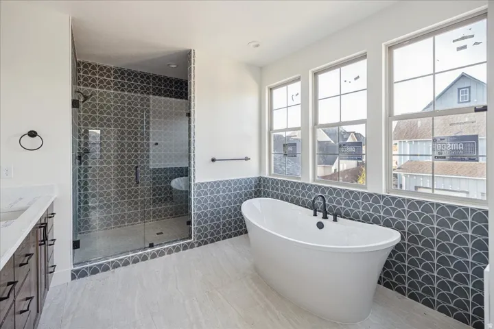 Freestanding soaking tub with matte black fixtures, glass-enclosed shower with decorative tile surround, dual vanity with quartz countertop, and large windows providing abundant natural light