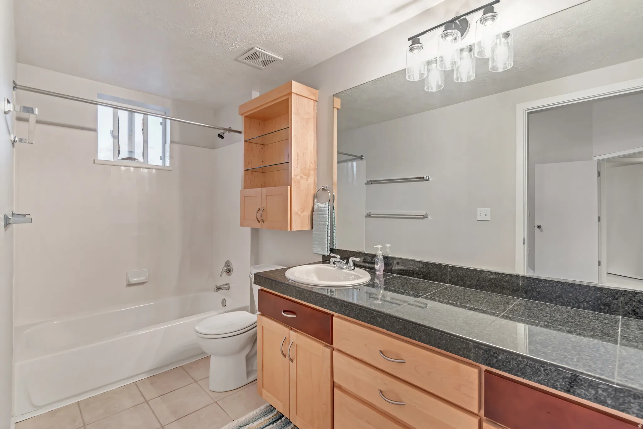 White drop-in bathtub with shower, light wood-finish cabinetry, dark stone-finish countertop, and a large vanity mirror