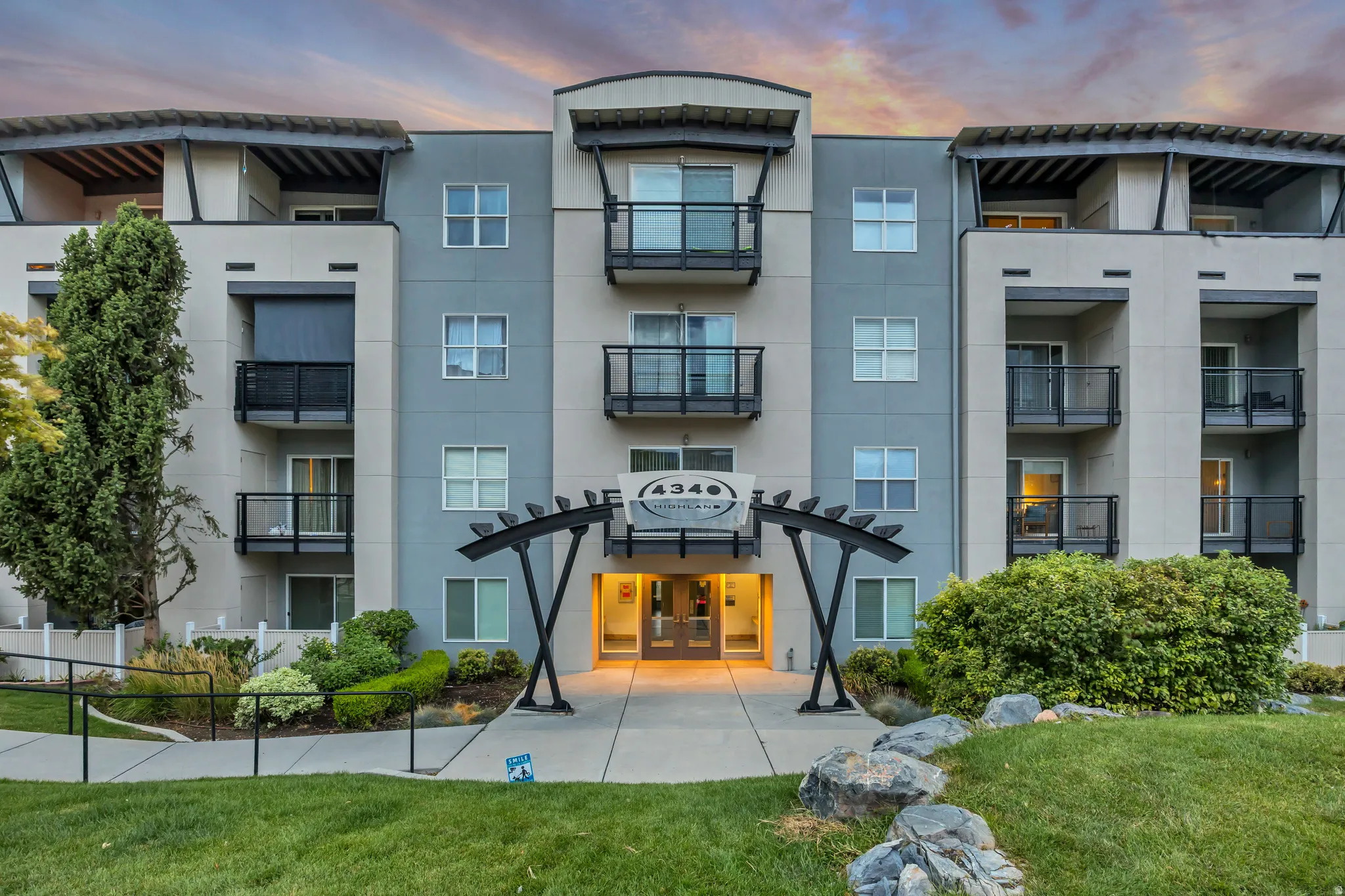 Multi-story building exterior with a modern facade, individual balconies with metal railings, a pergola entrance feature, manicured landscaping, and a paved walkway