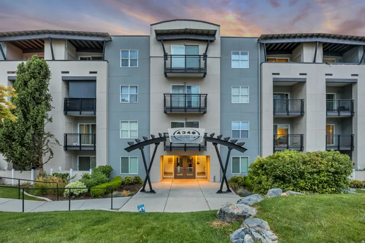 Multi-story building exterior with a modern facade, individual balconies with metal railings, a pergola entrance feature, manicured landscaping, and a paved walkway