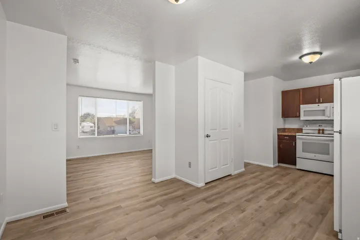 Open-concept layout with wood-finish flooring, white walls, kitchen with white appliances, dark wood-finish cabinetry, and a pantry with a white paneled door