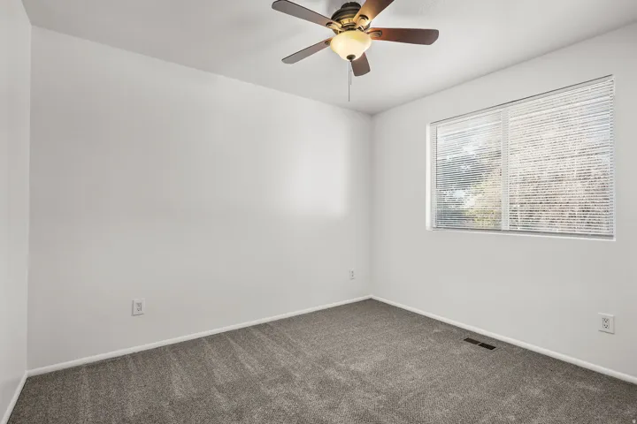 Neutral white wall paint, grey carpeting, ceiling fan with integrated lighting, window with horizontal blinds