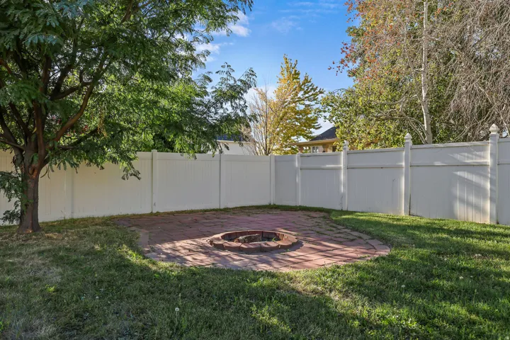 Expansive yard space featuring a paver patio and a built-in fire pit, surrounded by a white privacy fence and mature trees