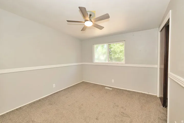 Ceiling fan with integrated lighting, window with exterior exposure, carpeted flooring, wall trim, and a built-in closet with dark wood-finish doors