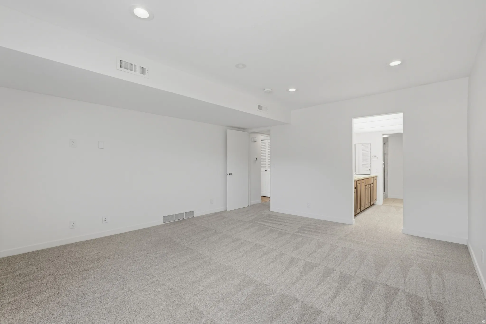 Recessed ceiling lighting illuminates a spacious room featuring light-colored patterned carpeting and crisp white walls