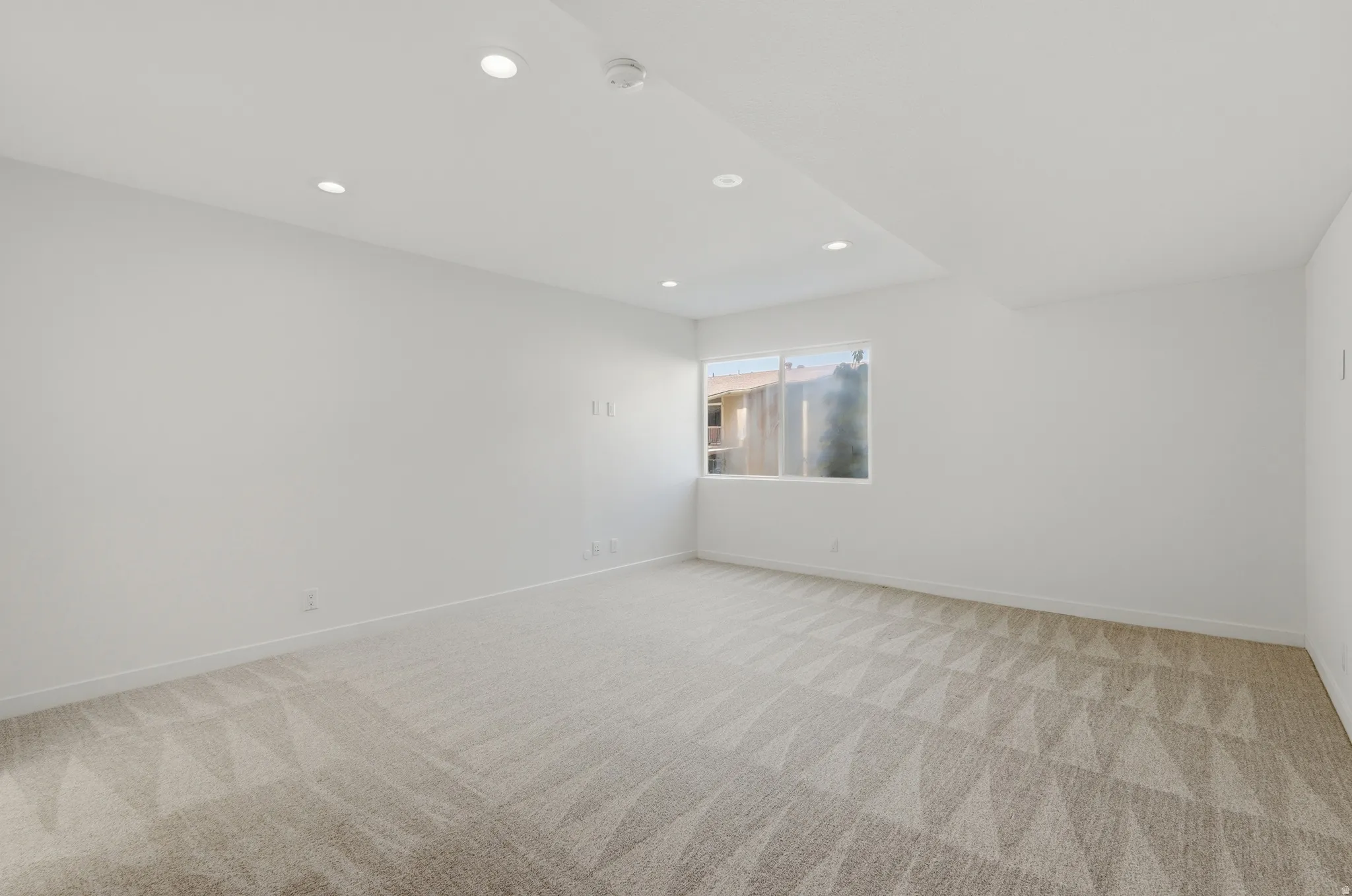 Recessed lighting illuminates a spacious room featuring a window and patterned carpeting
